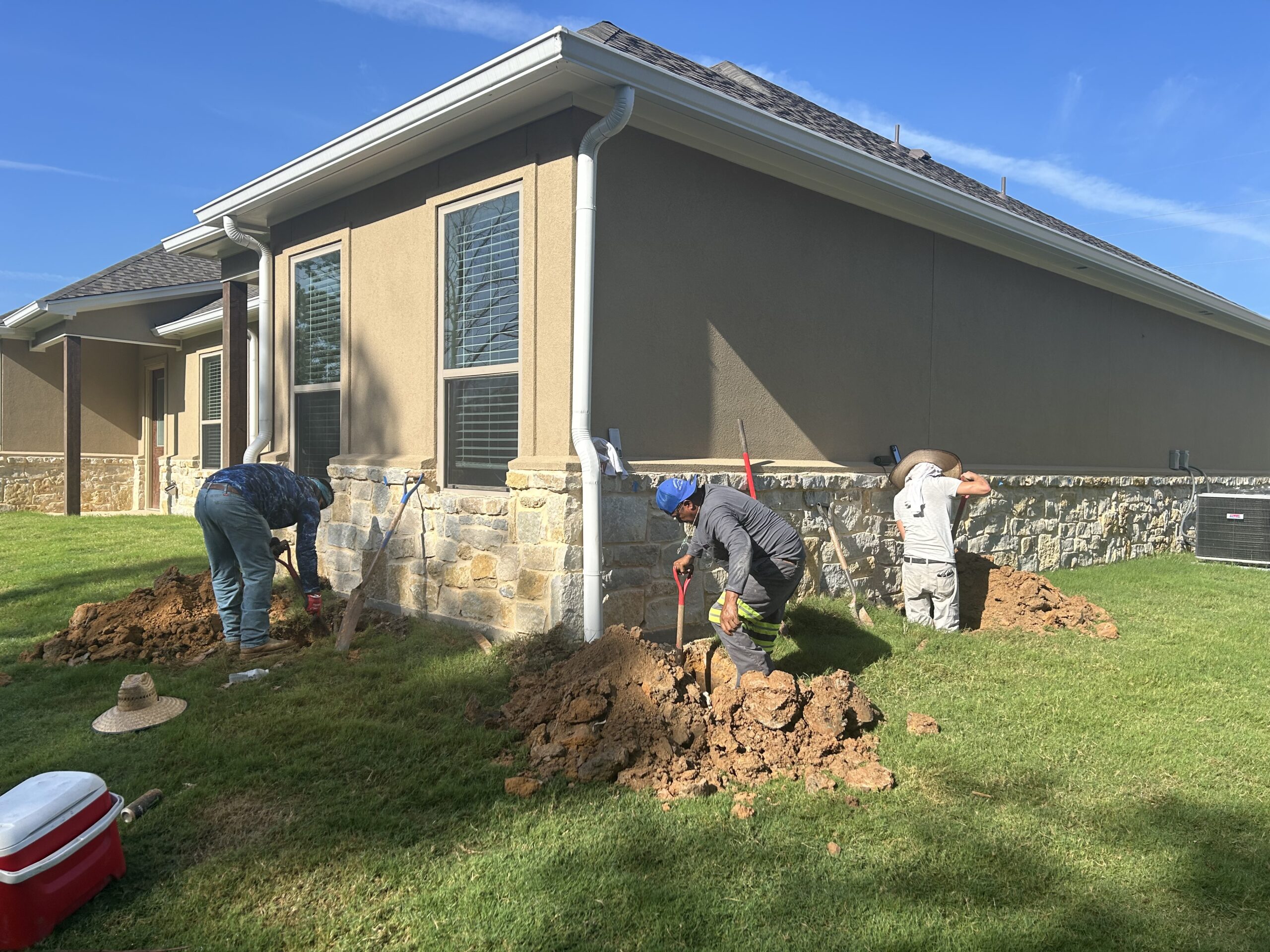 Foundation repair team digging trenches along the exterior wall of a stucco and stone home in East Texas.